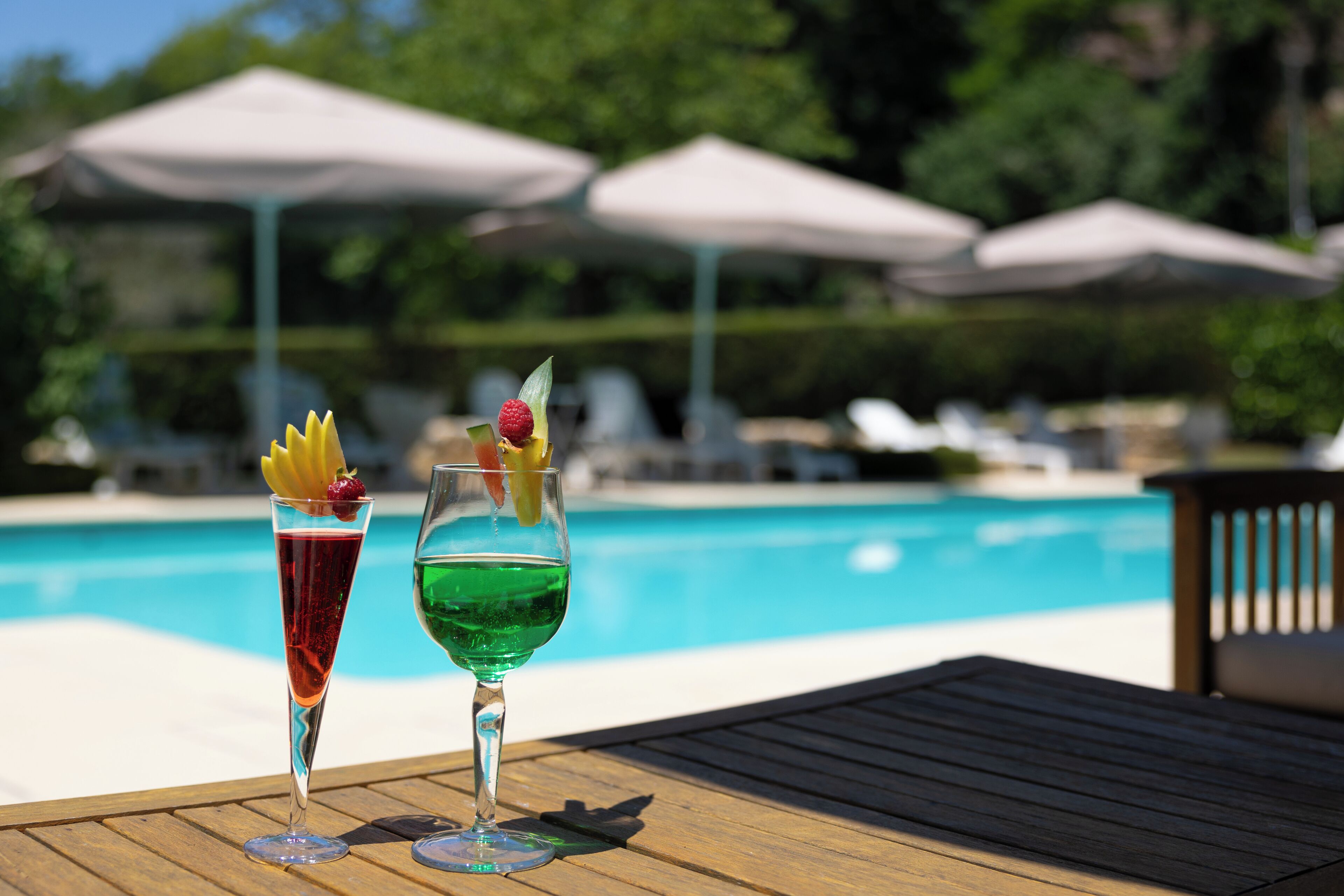 Two cocktails on a wooden table by a poolside with umbrellas and lounge chairs in the background.