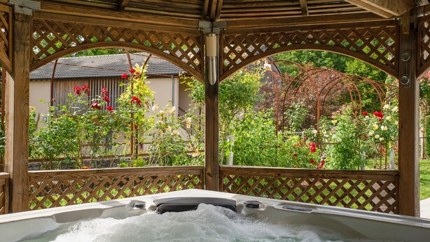 Uma banheira de hidromassagem sob uma pérgola de madeira com laterais treliçadas, cercada por vegetação exuberante e plantas floridas.