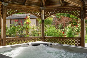 A hot tub under a wooden pergola with lattice sides, surrounded by lush greenery and flowering plants.