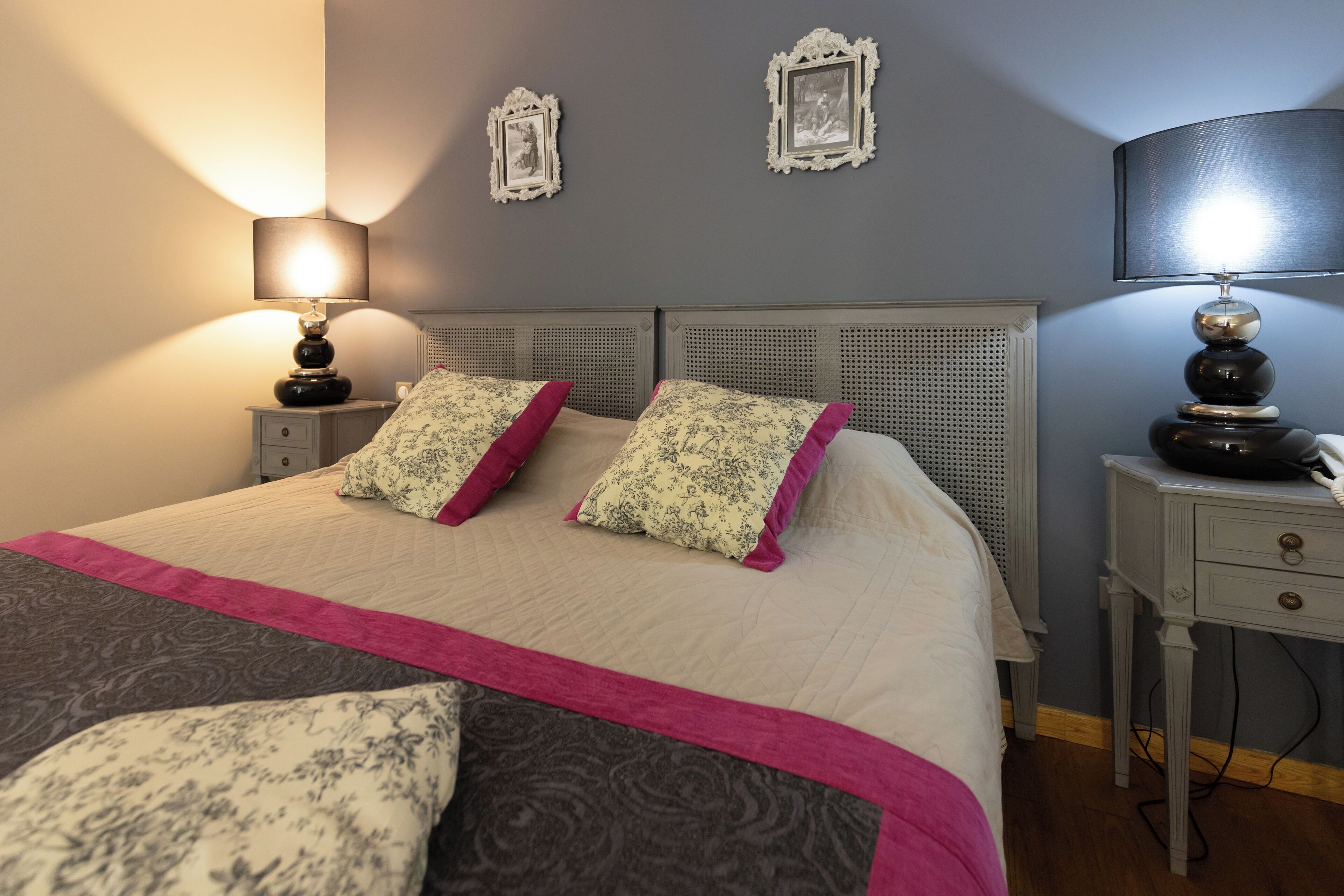 A neatly made bed with floral and solid pillows, a bedside table with a lamp, and two wall-mounted picture frames.
