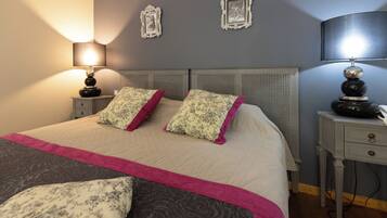Een opgemaakt bed met bloemen- en effen kussens, een nachtkastje met een lamp, en twee aan de muur bevestigde fotolijstjes.