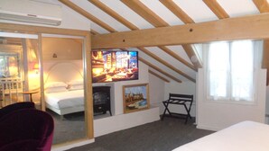 Quarto com cama, uma cadeira, um quadro, televisão e janela com cortinas.