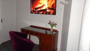 Um quarto com escrivaninha de madeira, uma cadeira roxa, uma TV na parede mostrando uma lareira, e um vaso com flores sobre a escrivaninha.