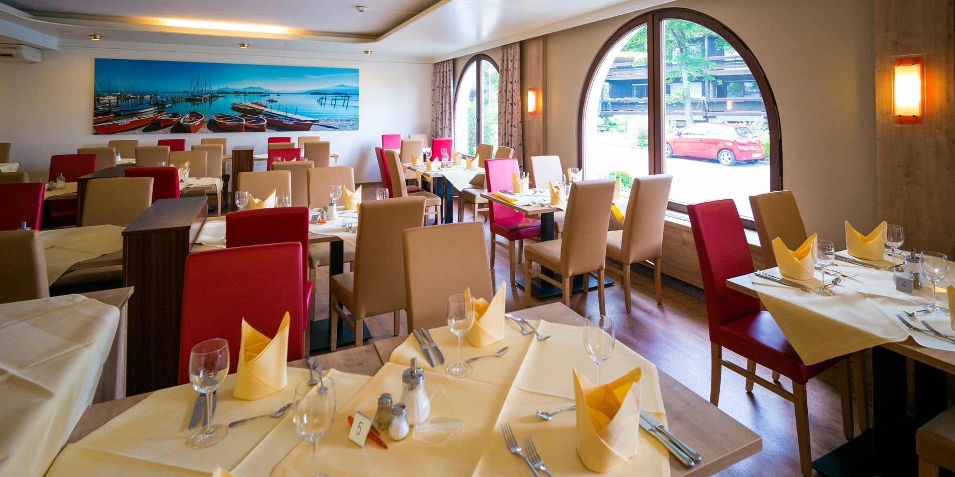 A restaurant with tables set for dining, chairs, and a painting of a harbor on the wall.