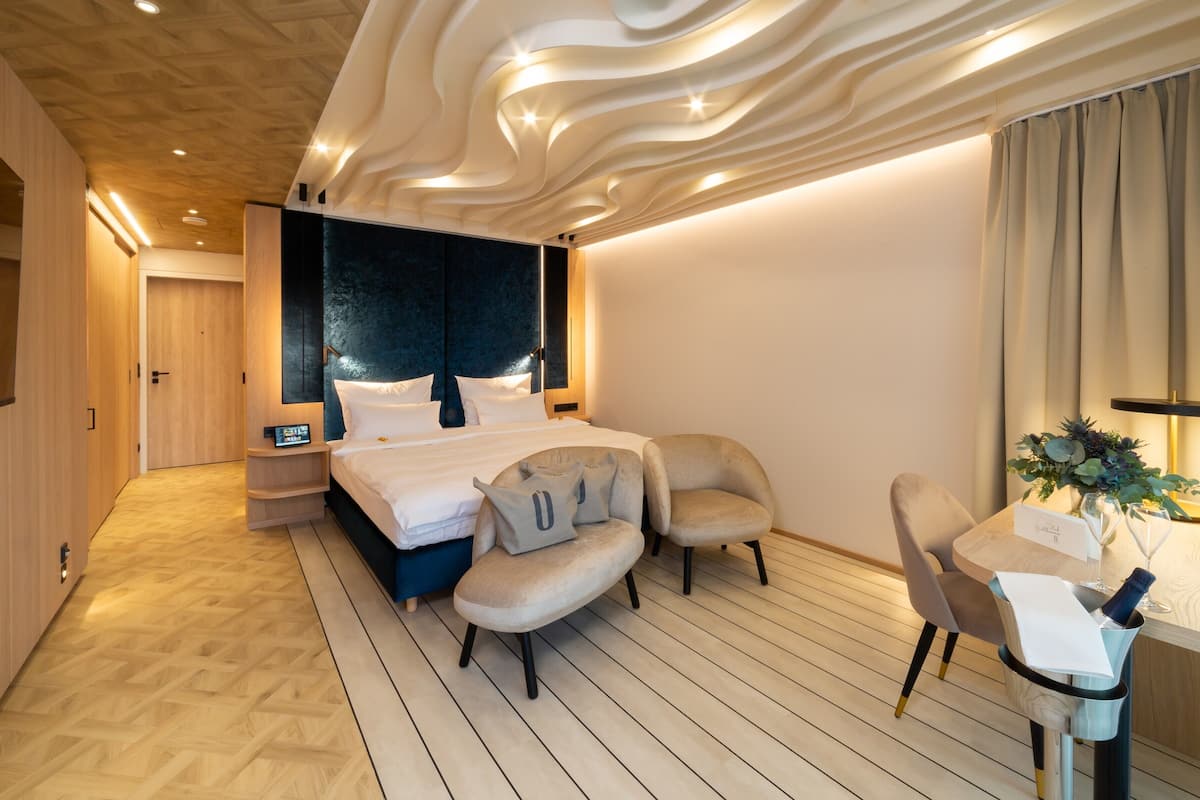 A modern hotel room with a wooden floor, a large bed with white linens, two armchairs, a small table with a vase and wine glasses, and a wall-mounted TV.