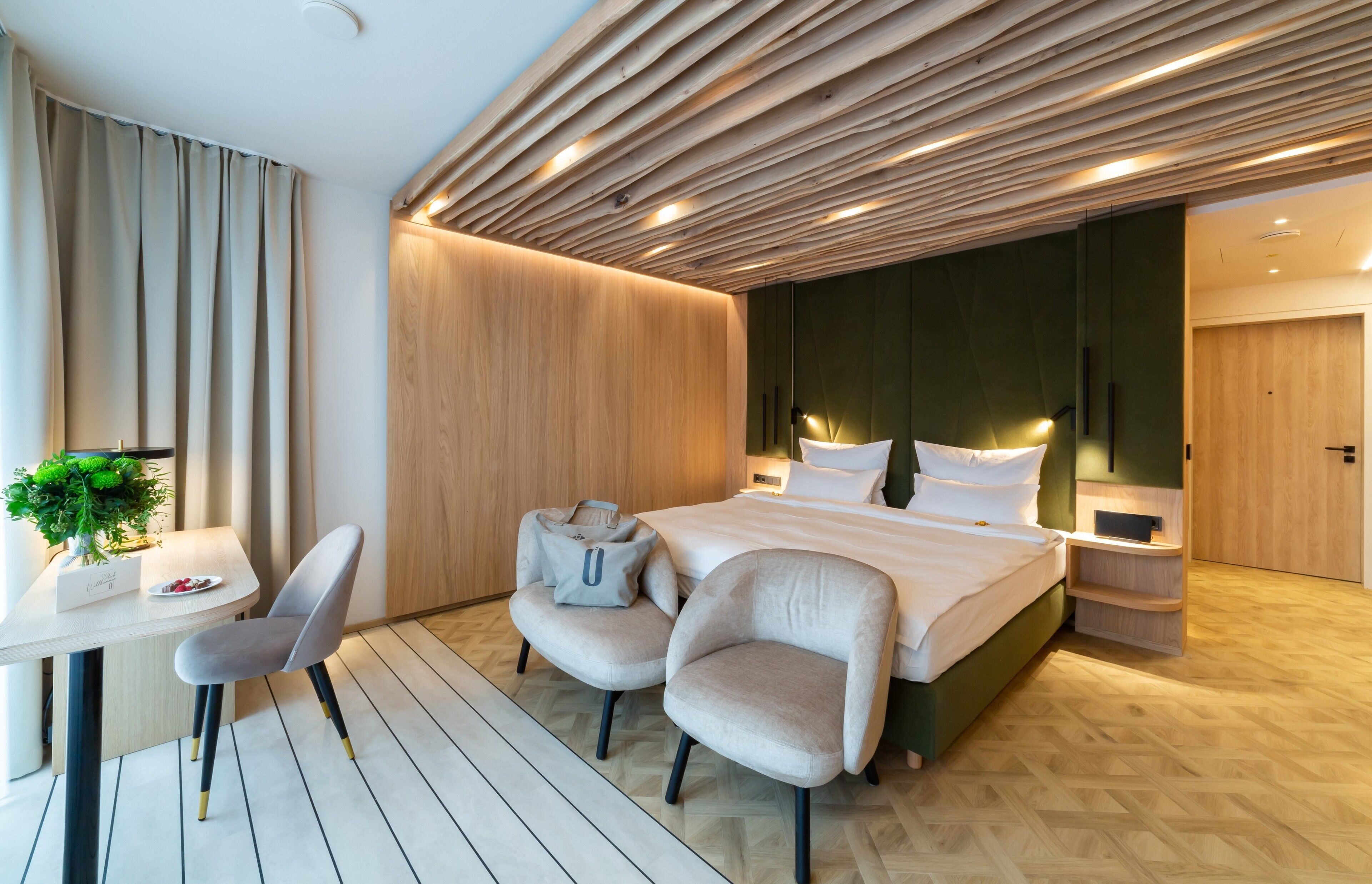 A modern hotel room with a wooden ceiling, a bed with white linens, two armchairs, a small table with a plant, and a bedside table with a lamp.