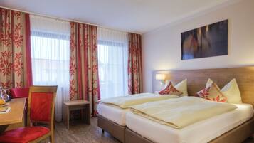 A hotel room with a bed, a desk with a chair, a small table with a chair, and a painting on the wall.