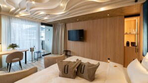 A modern hotel room with a large bed, a flat-screen TV mounted on the wall, a wooden cabinet, and a dining area with a table and chairs.