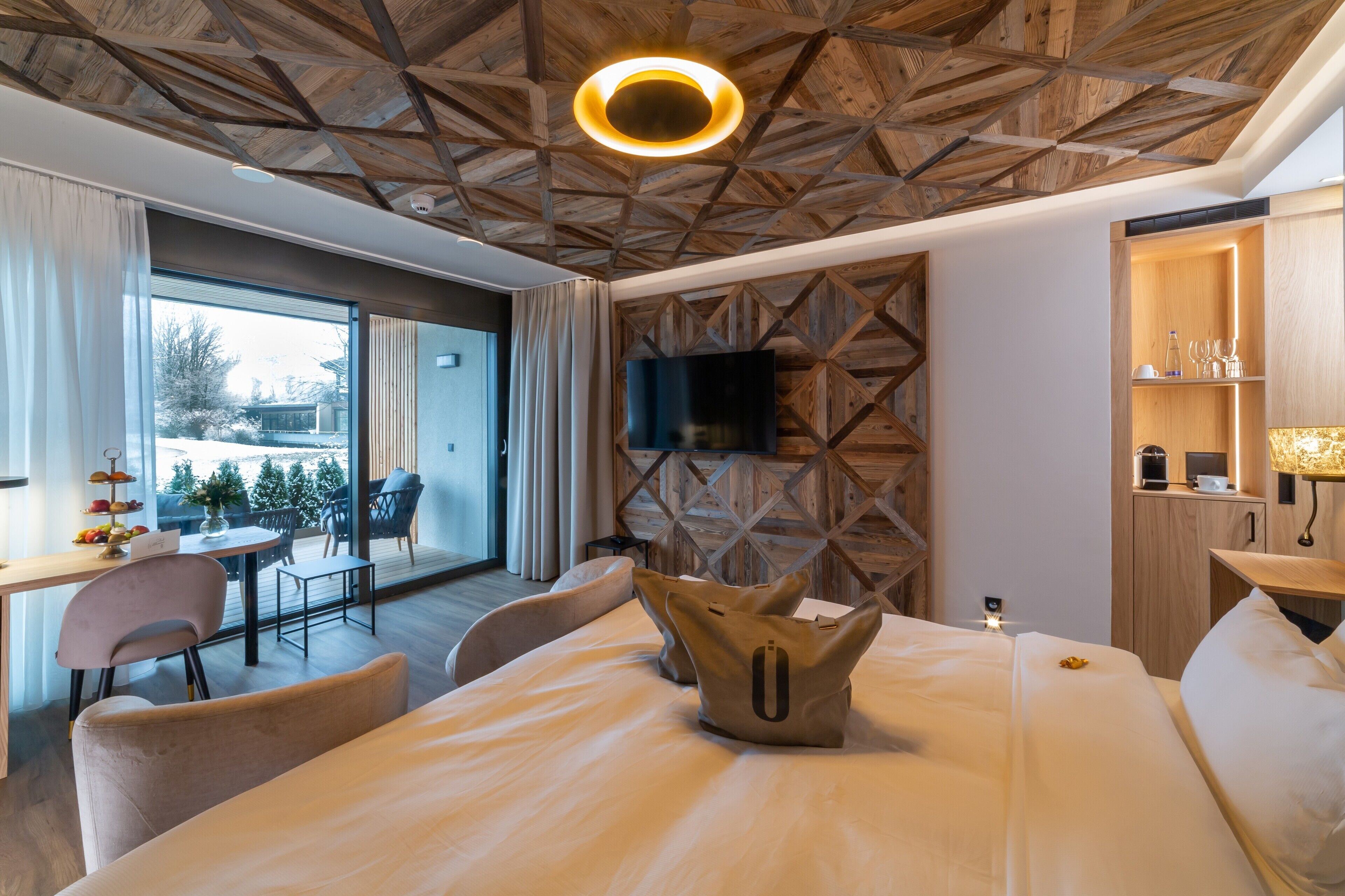 Una habitación de hotel moderna con techo de madera, una cama grande, un televisor, un escritorio con una silla y un balcón con mobiliario exterior.