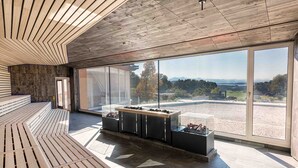 A spacious room with a wooden slatted ceiling, large windows offering a view of a gravel area and distant trees, and a seating area with a table and display cases.