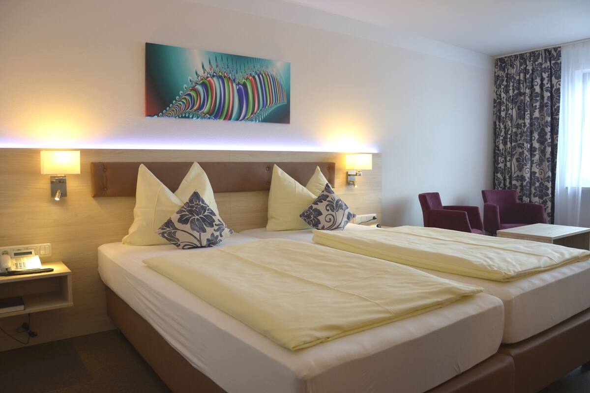 A hotel room with two beds, a desk, and a chair. There is a painting on the wall and a lamp on the nightstand.