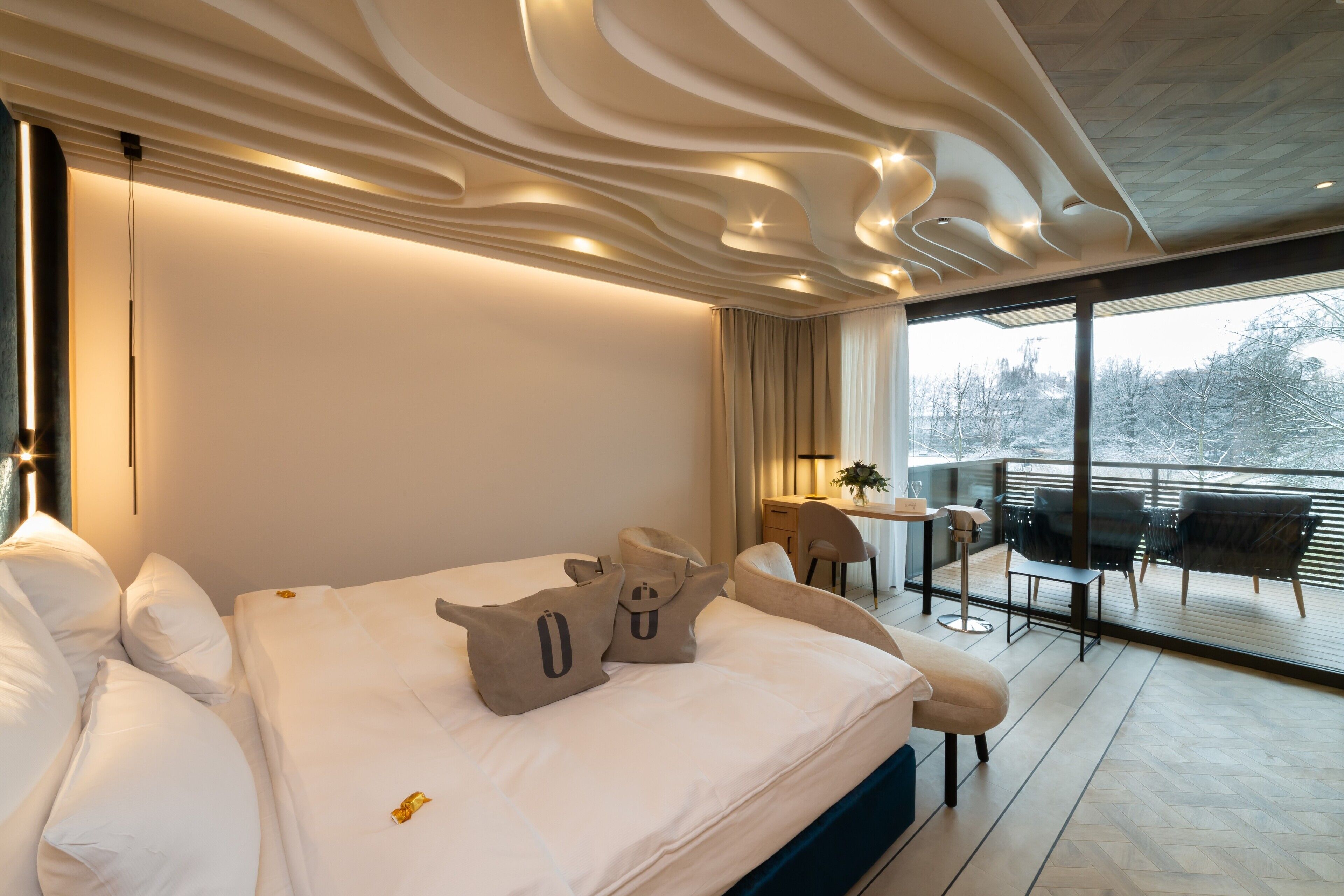 A modern hotel room with a large bed, a balcony with seating, and a view of trees.