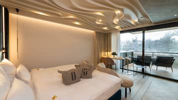 A modern hotel room with a large bed, a balcony with seating, and a view of trees.