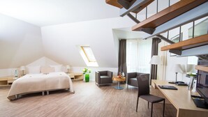 A modern hotel room with a bed, a desk, a chair, and a small table. There is a window with curtains and a skylight.