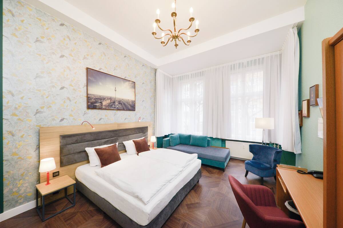 Ein Hotelzimmer mit einem großen Bett, einer Couch, einem Schreibtisch und einem Stuhl. Eine Kandelaber, ein Wandbild und ein Fenster mit Vorhängen sind ebenfalls vorhanden.