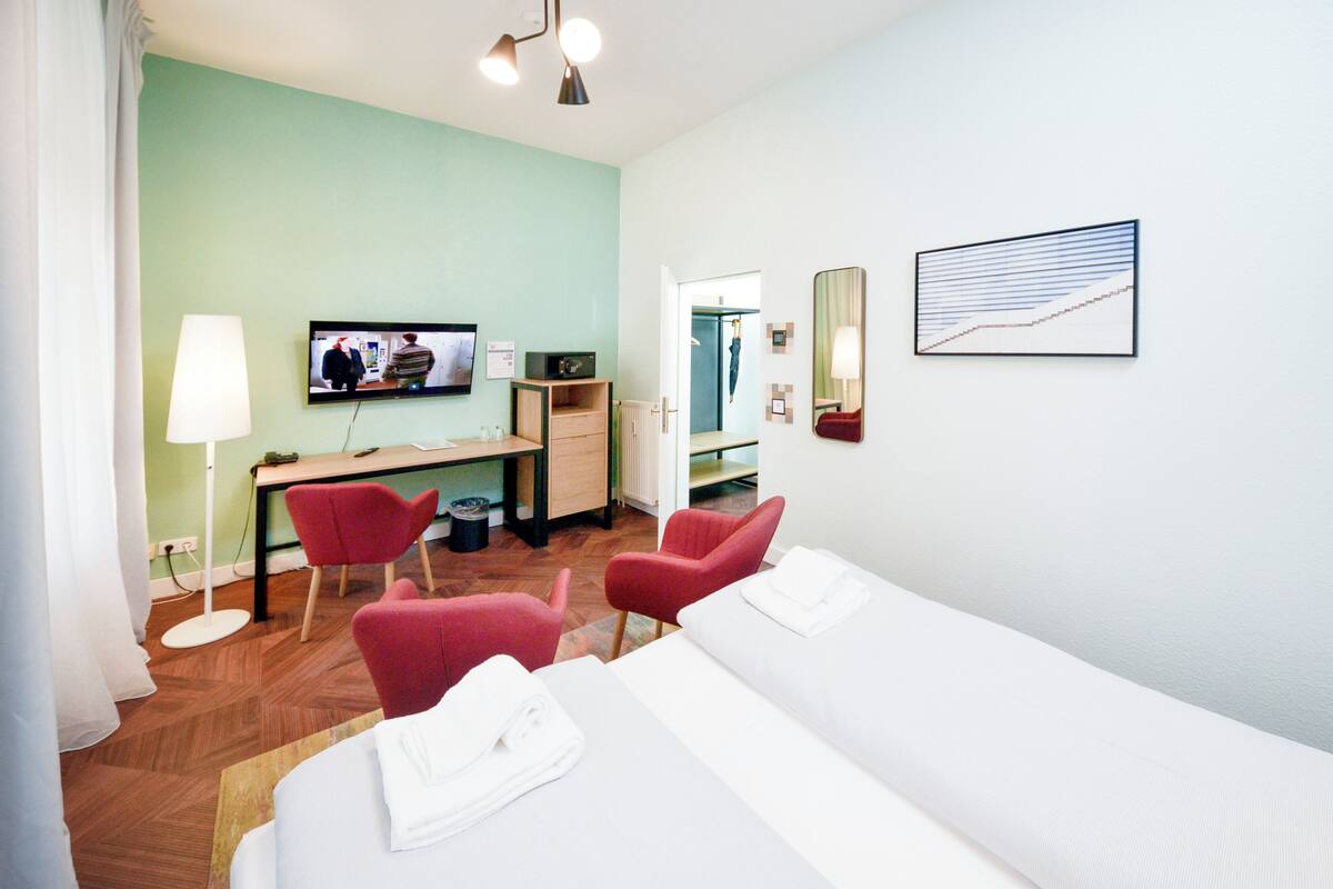 Ein Hotelzimmer mit einem Bett, zwei roten Stühlen, einem Schreibtisch mit Computer, einem Fernseher und einem Spiegel.