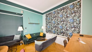 A modern living room with teal walls, a floral wallpaper, a dark green sofa, a grey armchair, a wooden dining table, and a small round table. - AMC Hotel - Schöneberg (Berlin)