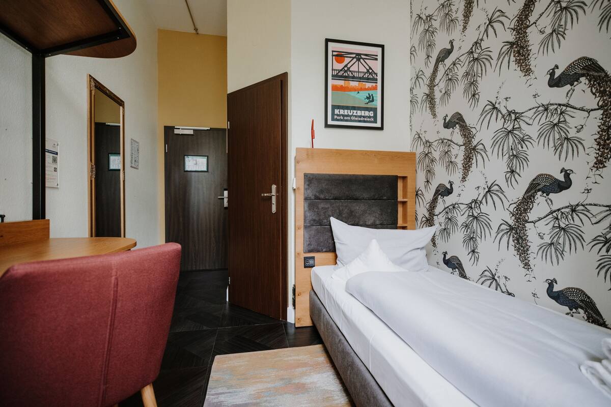 Ein Hotelzimmer mit einem hölzernen Bett, einem roten Sessel, einem Holztisch und einer Wand mit Vogelmuster.
