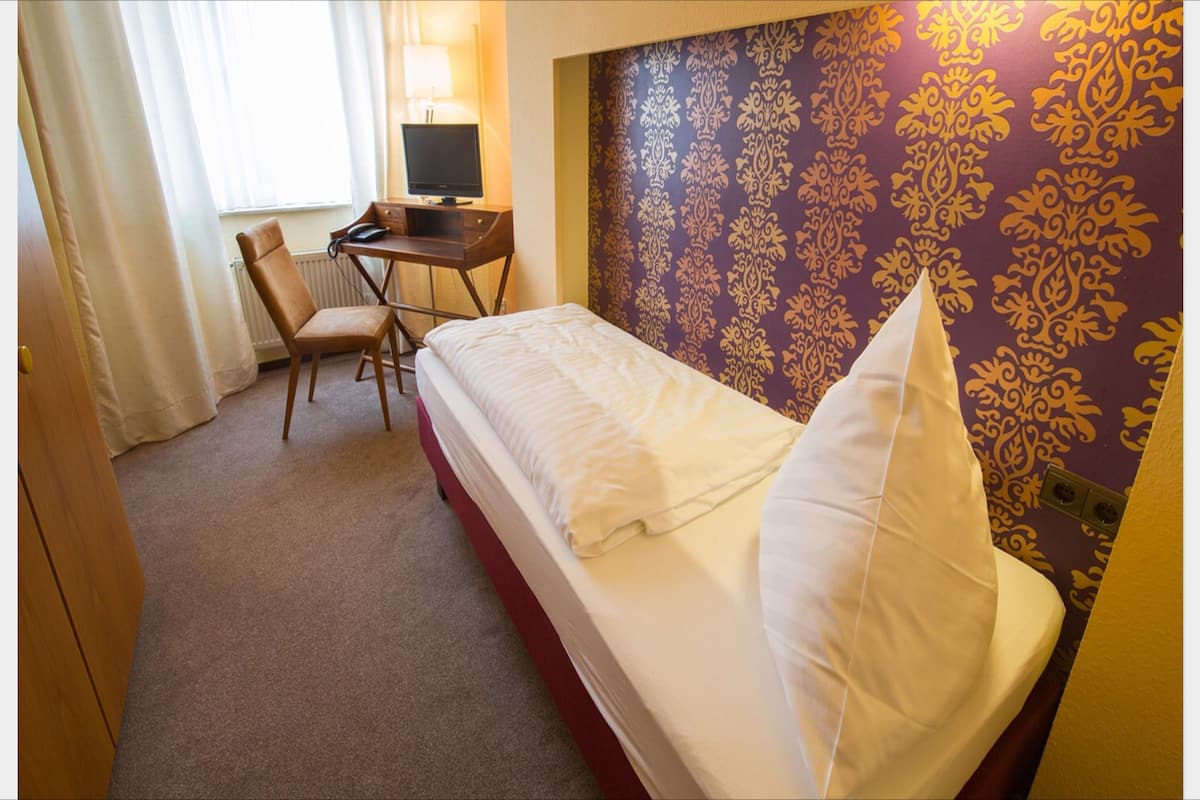 Ein Einzelbett mit weißer Bettwäsche, ein Schreibtisch mit Computer und Stuhl sowie eine Wand mit Muster.