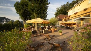 An outdoor seating area with wooden chairs and yellow umbrellas, surrounded by potted plants and a building with flower boxes. - BSW - Hotel Lindenbach (Nievern)