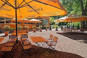 An outdoor seating area with wooden tables and chairs, covered by large orange umbrellas, and surrounded by potted plants.