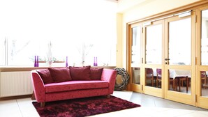 A living room with a pink sofa, a dark rug, and a view through a glass door to an outdoor dining area. - BSW - Hotel Lindenbach (Nievern)
