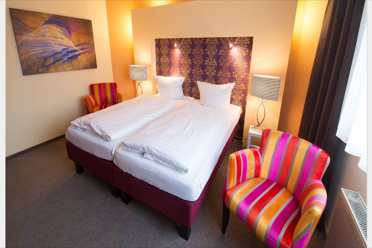 Ein Hotelzimmer mit einem großen Bett, zwei Nachttischen, einem farbenfrohen Streifenstuhl und einem dekorativen Kopfteil.