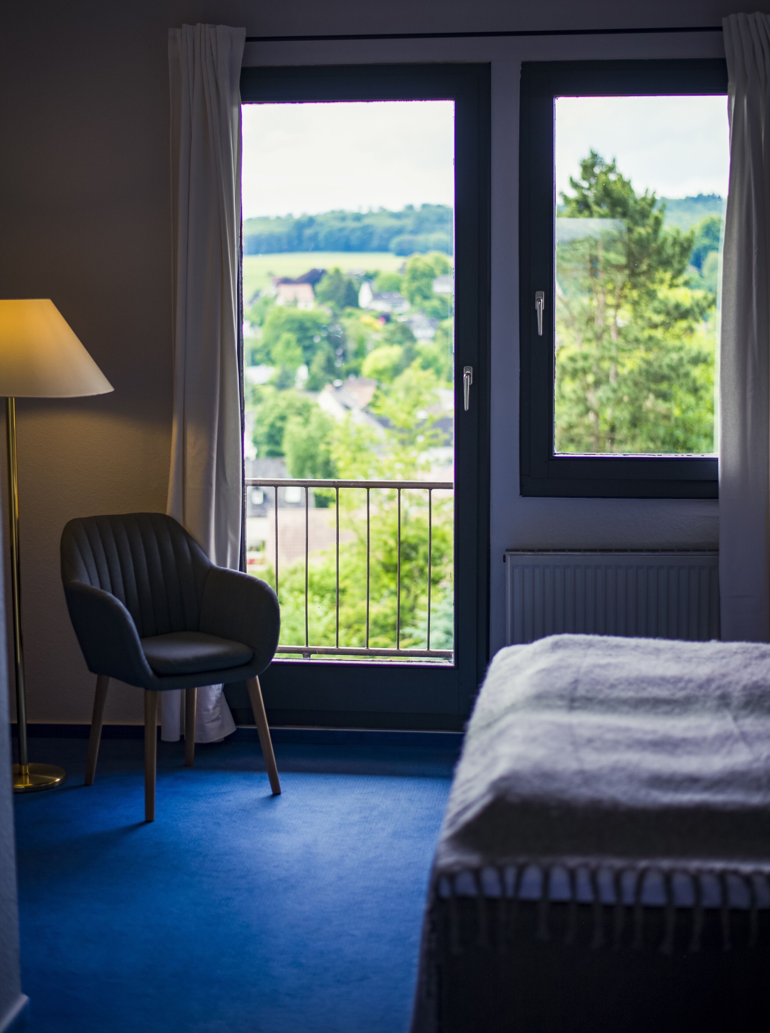 Ein Hotelzimmer mit Bett, Stuhl, Fenster und Blick ins Grüne sowie einer Lampe.