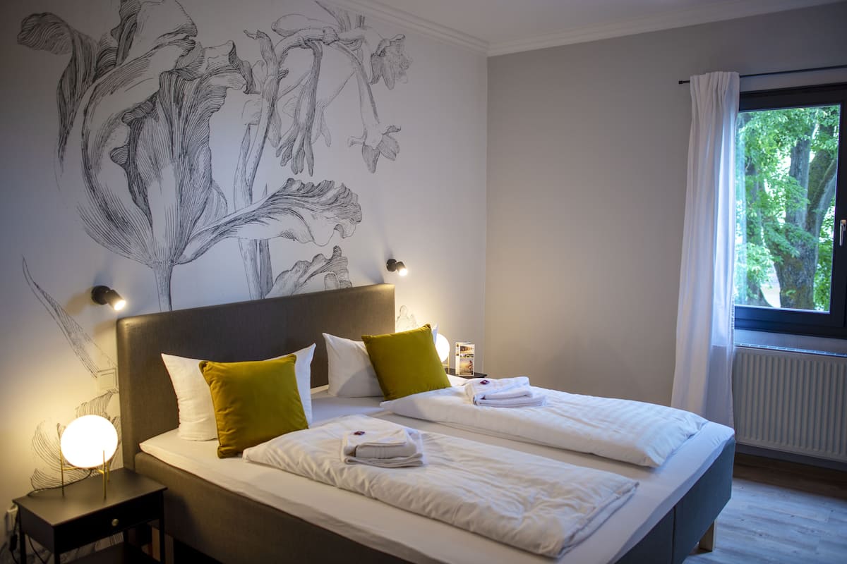 Habitación de hotel con dos camas, un mural grande en la pared, una mesita de noche con lámpara y una ventana con vista a follaje.