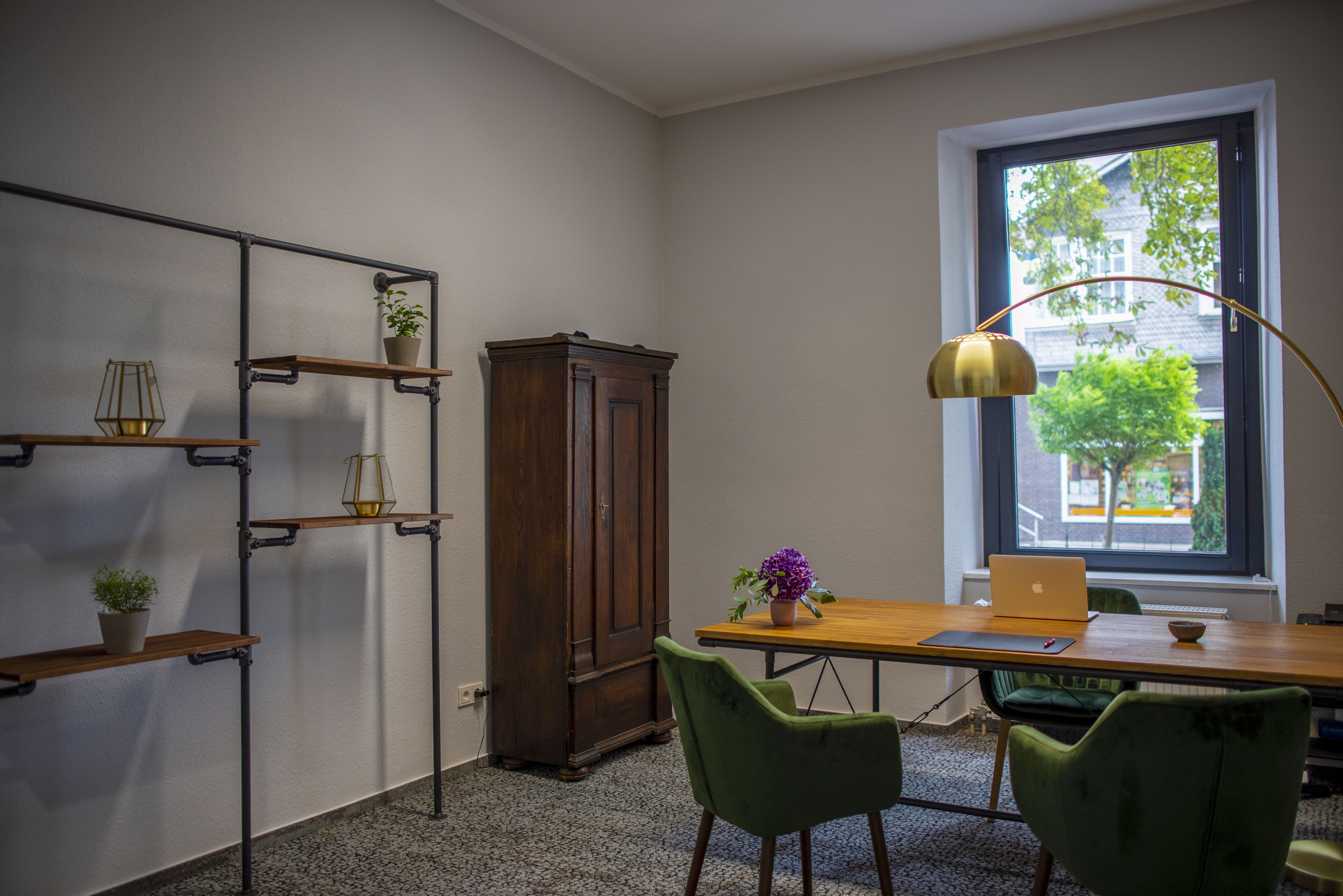 A room with a wooden desk, two green chairs, a laptop, a lamp, a cabinet, and shelves with plants.