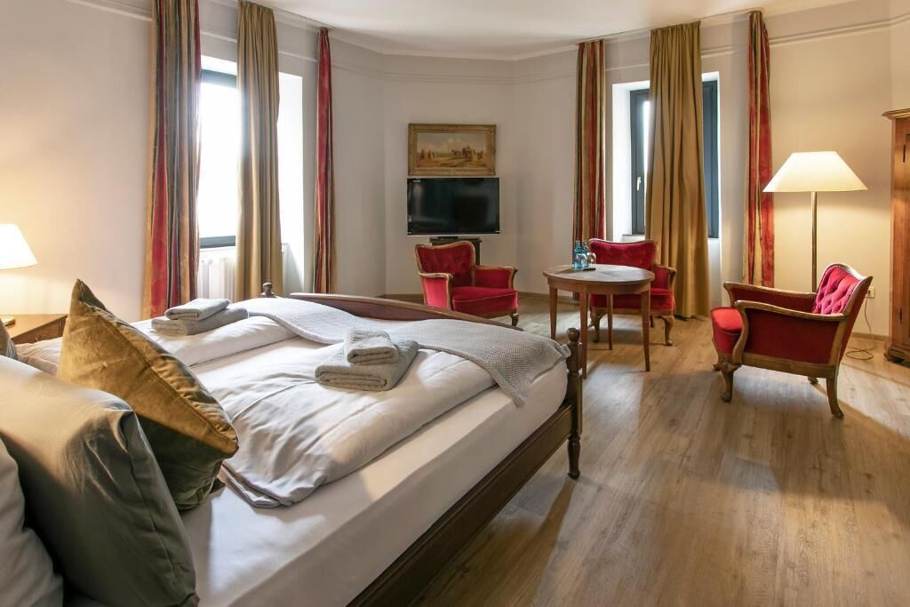 Ein Hotelzimmer mit einem Bett, zwei roten Sesseln, einem kleinen Tisch, einem Fernseher und einem Gemälde an der Wand.
