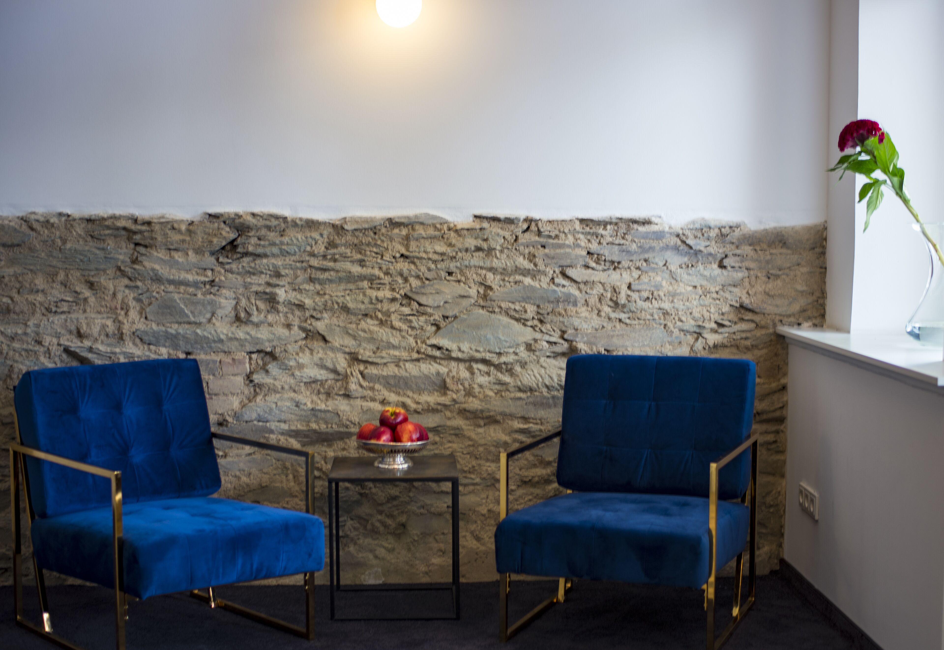A room with two blue chairs, a small table with a fruit bowl, a stone wall, and a vase with a flower on a windowsill.