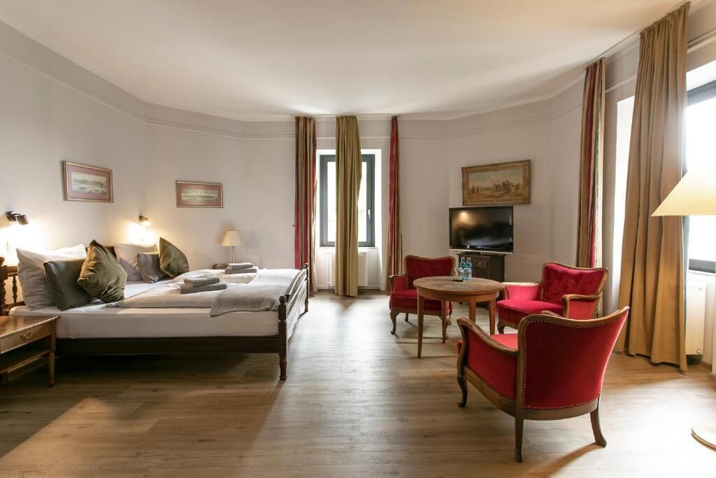 A hotel room with a bed, two red armchairs, a small table, a television, and a painting on the wall.