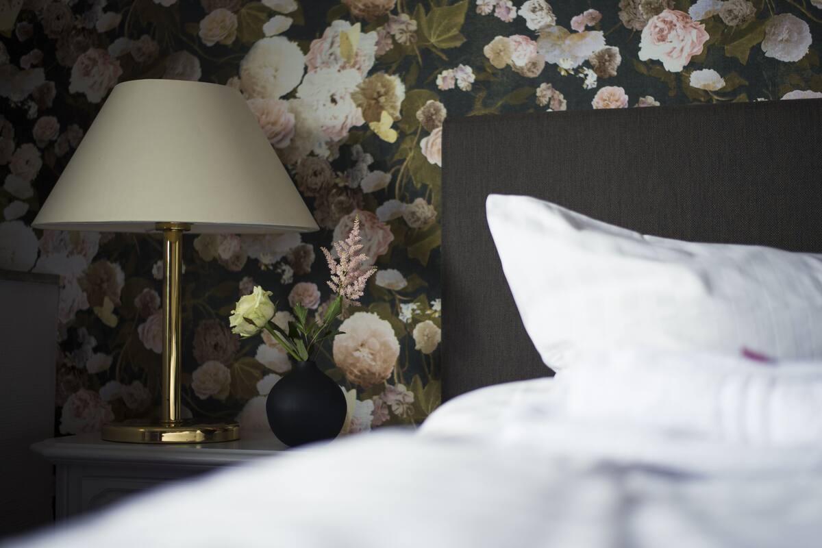 Una cama con ropa de cama blanca, un florero negro con flores y una lámpara en una mesita de noche.