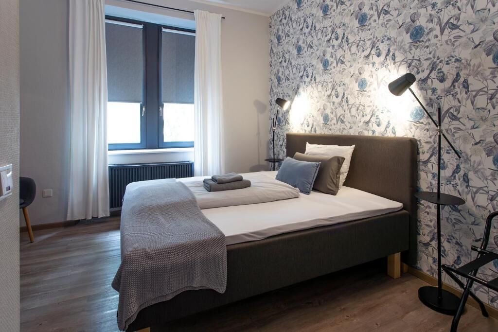 Ein Schlafzimmer mit einem Bett, einem Fenster mit Vorhängen, einem Heizkörper und einer Wand mit einem Blumenmuster.