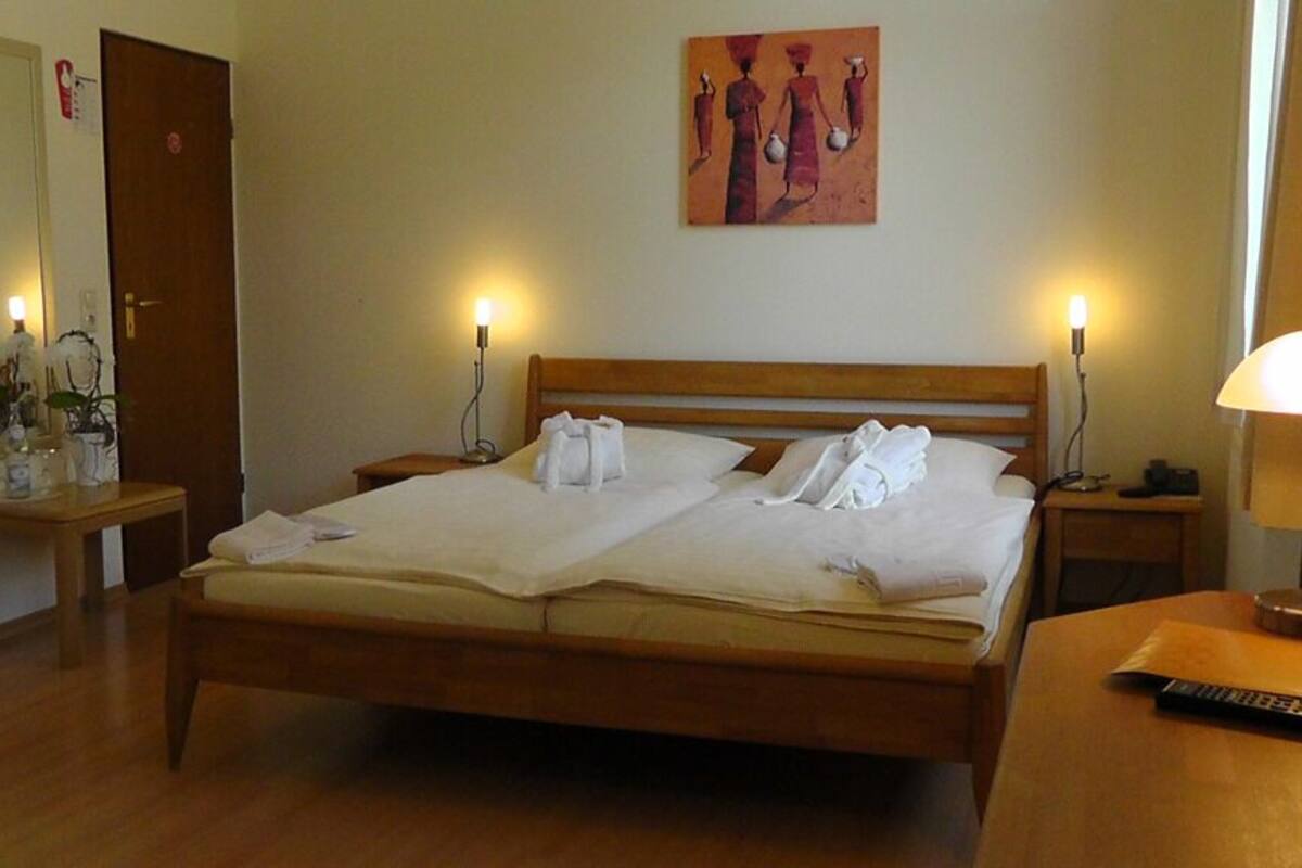 A neatly made bed with pillows and towels, a bedside table with a lamp, a remote control, and a painting on the wall.