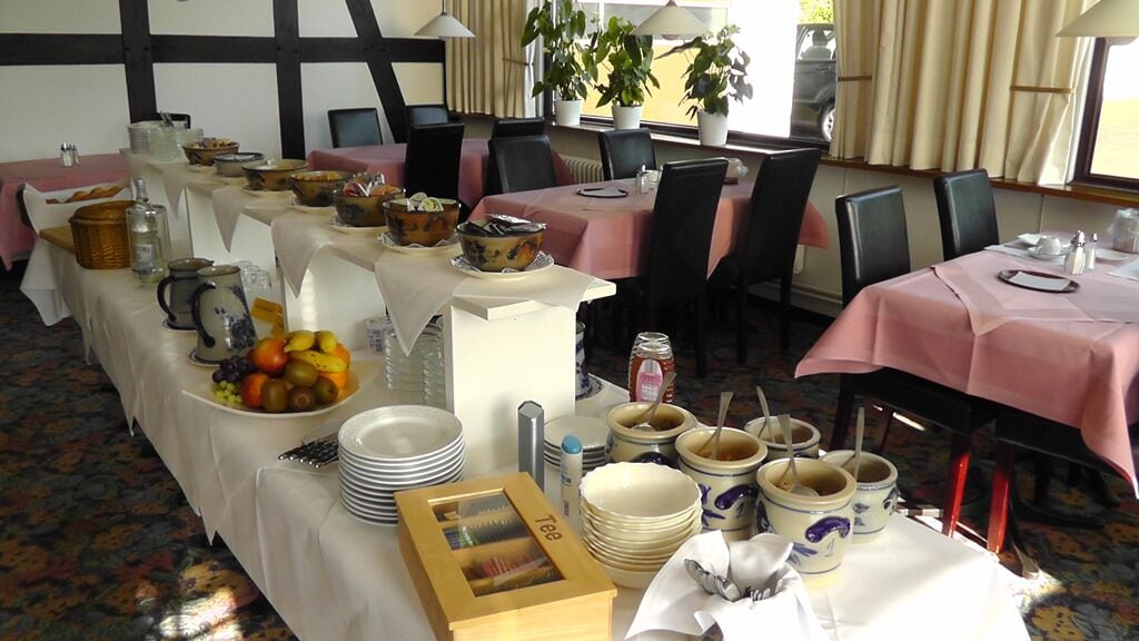 Une salle à manger avec plusieurs tables préparées pour le petit-déjeuner, chacune recouverte d’une nappe rose. On y trouve différents plats et ustensiles, et des plantes en pot sont disposées près des fenêtres.