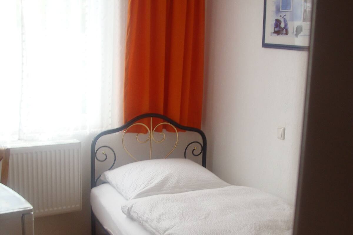 Ein kleines Zimmer mit einem Einzelbett, Holzboden, einem Fenster mit orangefarbenen Vorhängen und einem weißen Heizkörper.