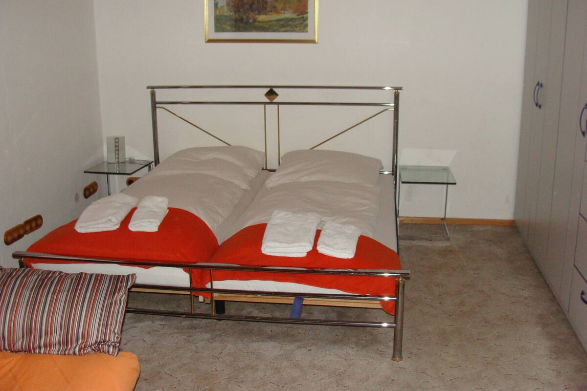 Ein Zimmer mit einem Einzelbett, Metallgestell, zwei Kissen und einer gestreiften Kopfbedeckung sowie einer roten Decke.