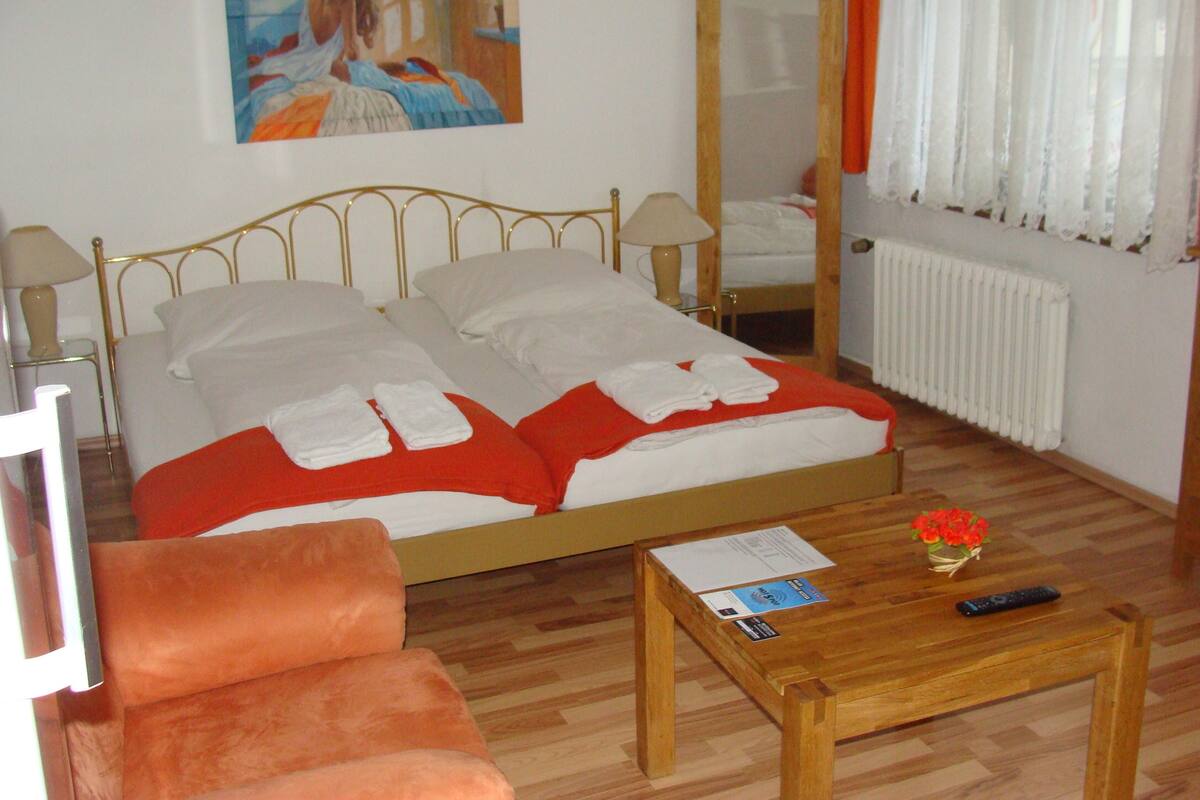 Ein Hotelzimmer mit einem Bett, einem Nachttisch aus Holz, einem Stuhl, einem Holztisch, einer Vase mit Blumen, einem Bild an der Wand und einem Fenster mit Vorhängen.