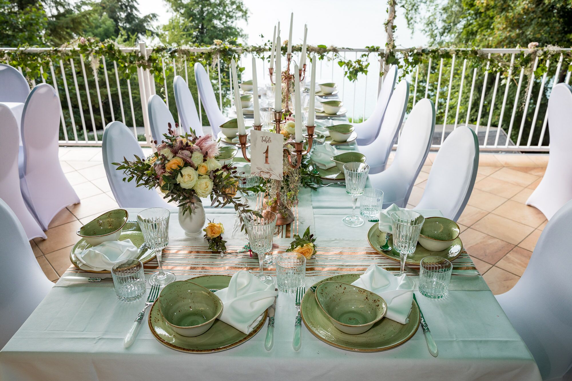 A long dining table set for a meal outdoors, surrounded by white chairs, with a floral centerpiece and candles.