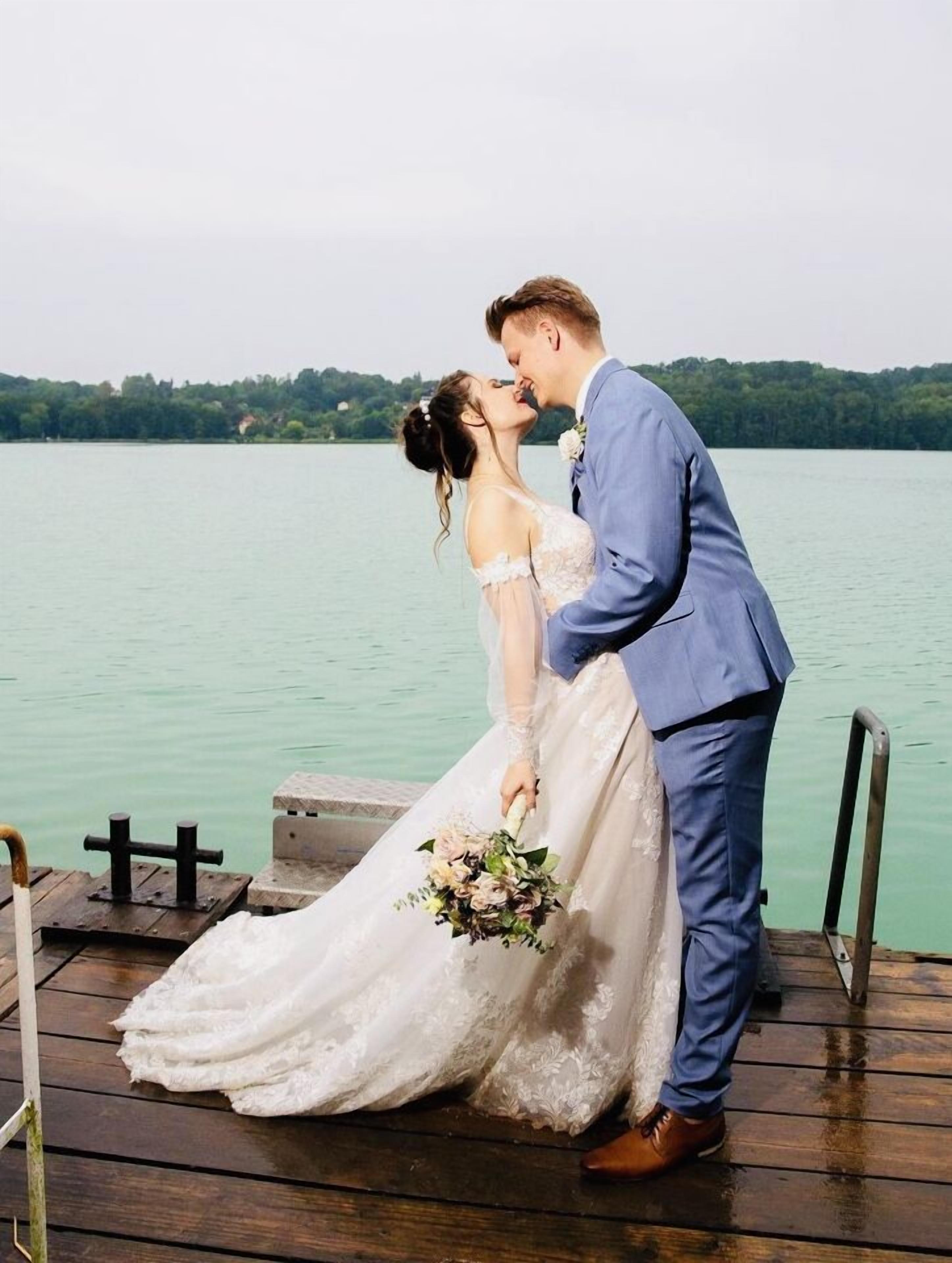 A couple on a wooden dock by a lake, with a wedding dress and a bouquet.