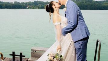 A couple on a wooden dock by a lake, with a wedding dress and a bouquet.