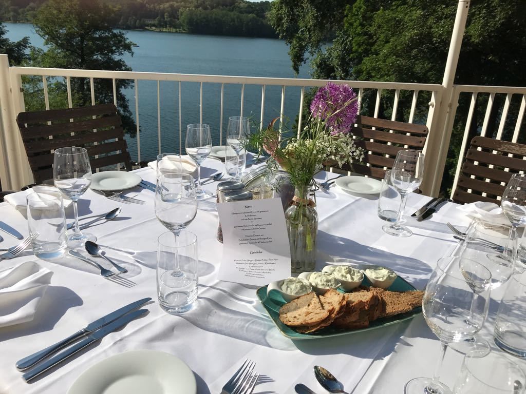 Et bord dækket til et måltid med menu, glas, bestik og et fad med brød og dyppelse, med udsigt over en sø.