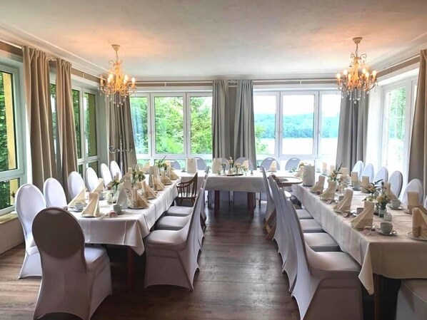 Un comedor elegante con mesas largas dispuestas para una comida, ventanas amplias y candelabros.