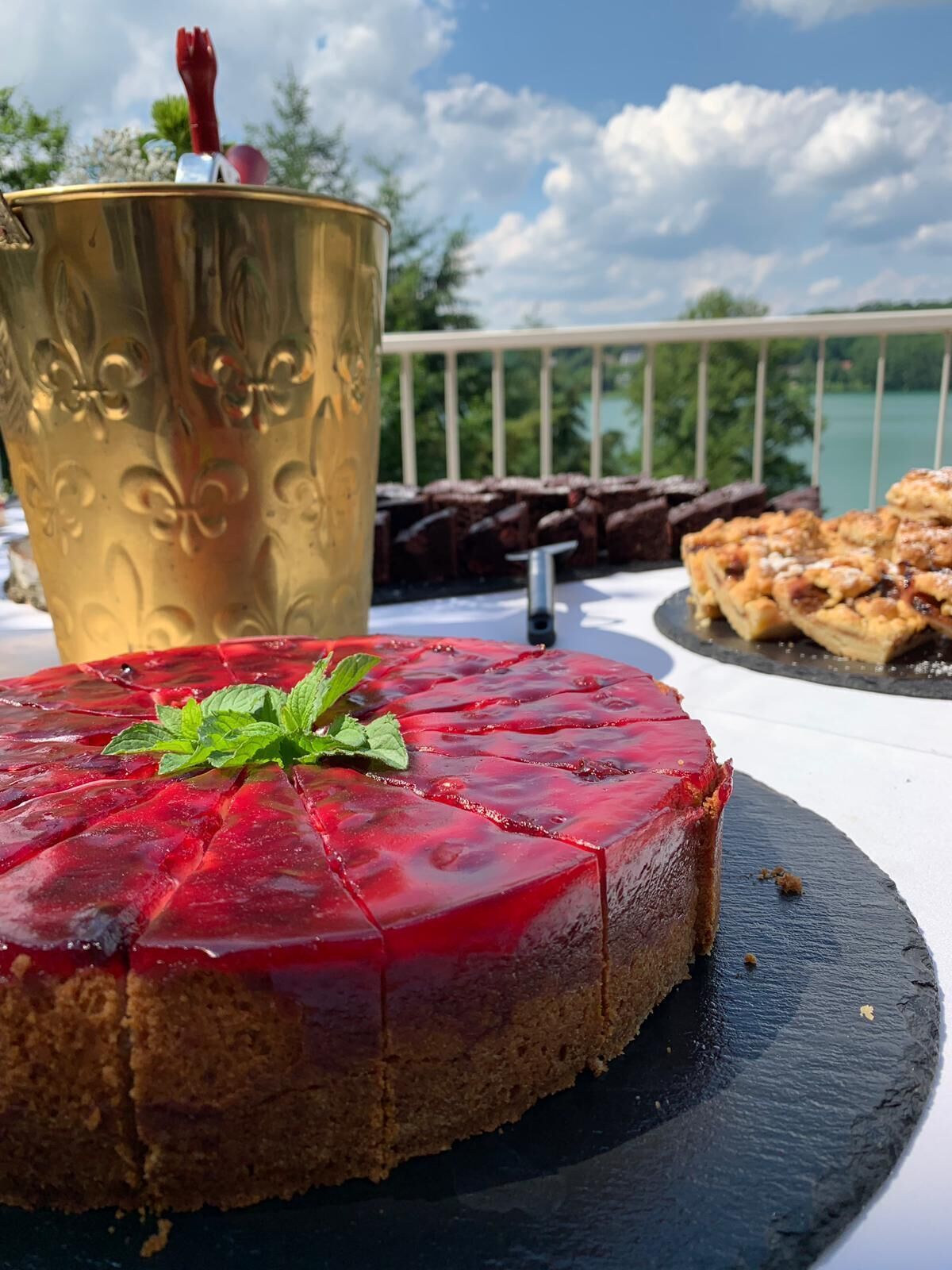 Un plato de postres con un pastel de remolacha, una taza dorada y una porción de tarta en un balcón con vistas a árboles y una masa de agua.