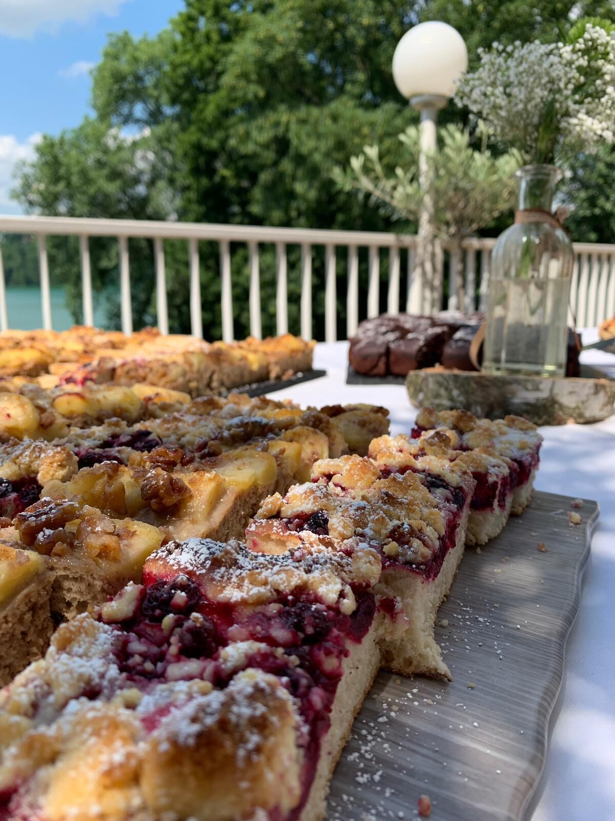 Una mesa con diversos postres, incluyendo un crumble de frutas, en una terraza al aire libre con barandilla y árboles de fondo.