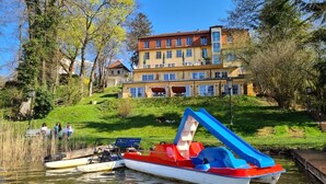 Un hôtel situé au bord de la rivière, avec un bateau amarré devant, une colline verdoyante avec un bâtiment en arrière-plan et un ciel dégagé.