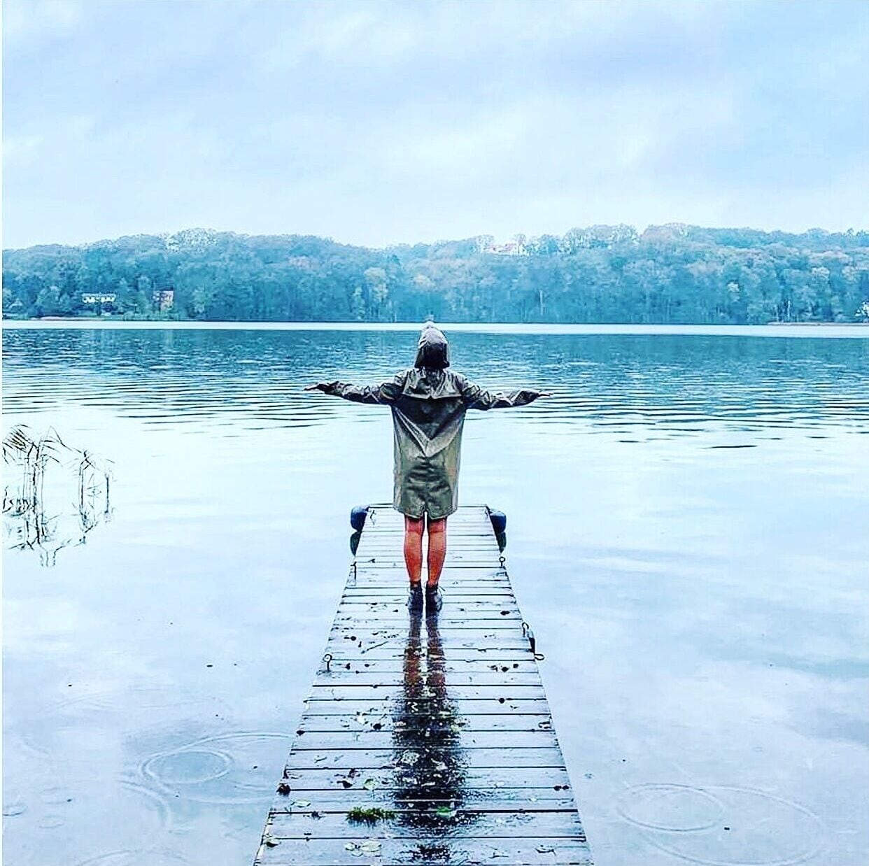 A person standing on a wooden dock extending into a body of water, with a forested hill in the background.