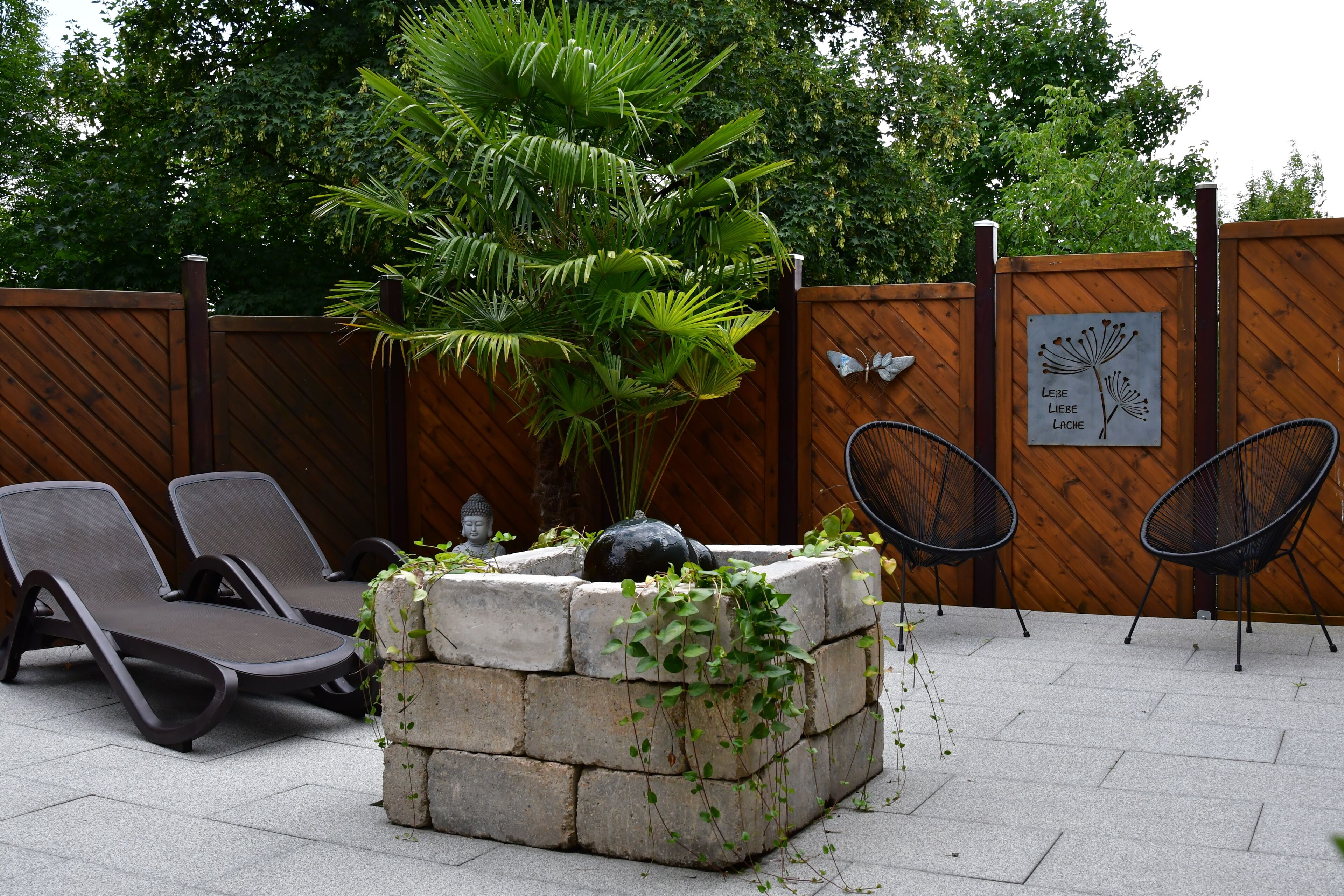 A patio area with lounge chairs, a stone planter with greenery, a Buddha statue, and a wooden fence.